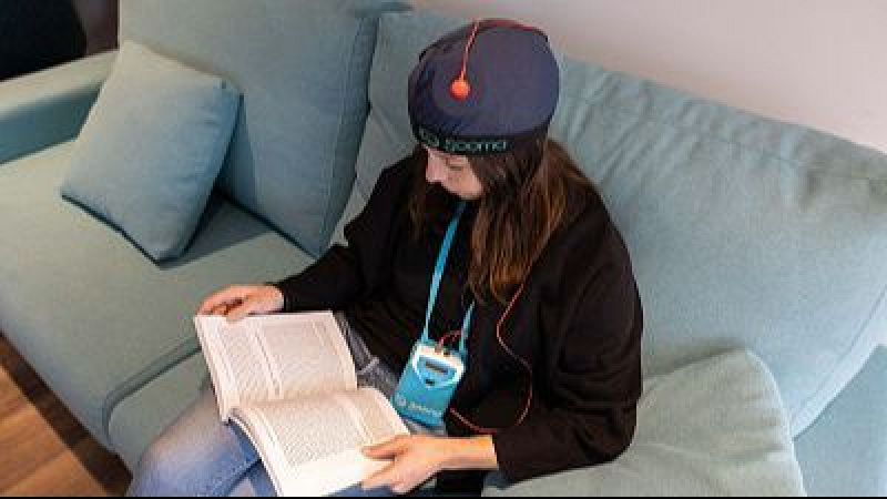 Mujer joven sentada en un sof� azul leyendo un libro mientras recibe una sesi�n de estimulaci�n cerebral con un dispositivo m�dico port�til.