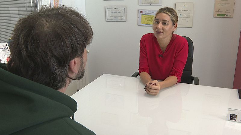 Dos personas conversan en un despacho con diplomas en la pared; una mujer con jersey rojo y coleta escucha atentamente a un hombre de espaldas sentado frente a ella.