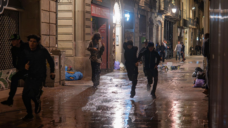 J�venes corriendo por una calle peatonal nocturna en Barcelona con suelo mojado y edificios cl�sicos con balcones de forja.