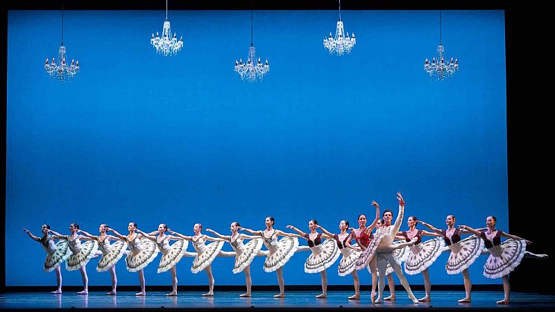 Bailarines de ballet cl�sico sobre un escenario con fondo azul y l�mparas colgantes, formando una simetr�a con tut�s blancos y detalles burdeos.