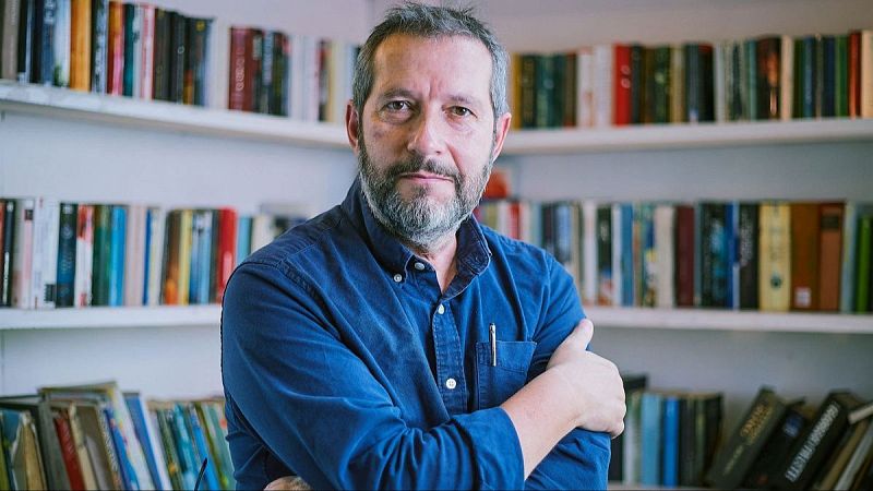 Retrato en plano medio del periodista Carles Porta con camisa azul marino y brazos cruzados en un estudio con libros de fondo.