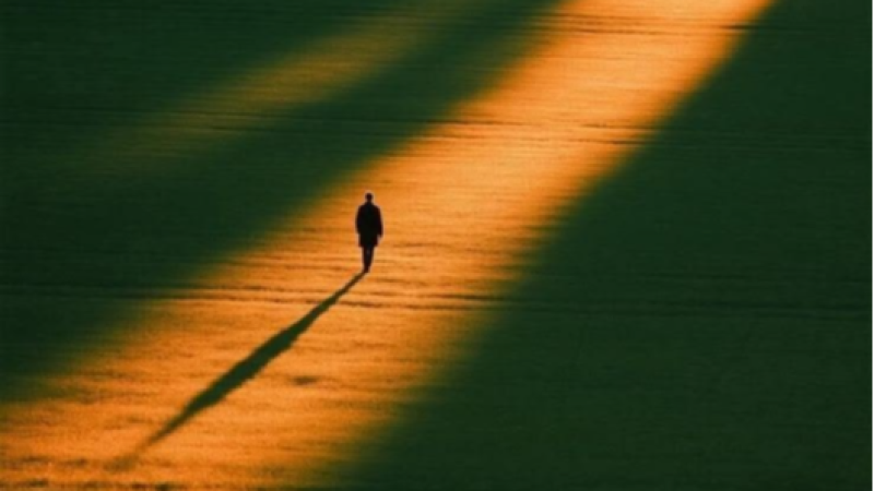 Imagen minimalista con un haz de luz dorada cruzando un campo oscuro y la silueta de una persona proyectando una sombra alargada.