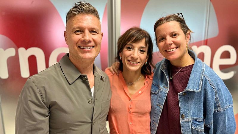 Tres personas sonríen de pie en un estudio de radio frente a paneles rojizos con logotipos circulares.