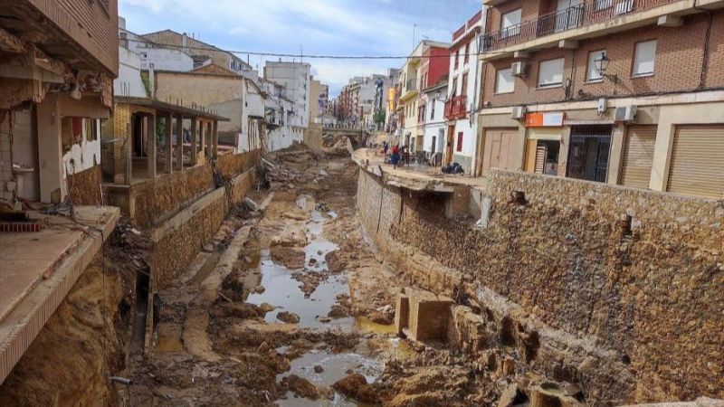 Graves destrozos en una localidad valenciana tras el paso de una riada, con viviendas parcialmente colapsadas, muros de contención socavados y el lecho del barranco anegado de lodo.