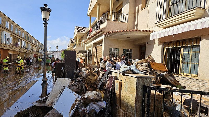Calle residencial inundada de lodo con vecinos, servicios de emergencia y escombros acumulados tras un temporal.