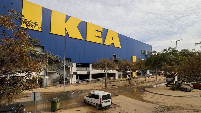 Fachada de un gran edificio comercial con letras amarillas, con los accesos y vehículos en la zona exterior completamente cubiertos de barro y lodo tras las inundaciones.