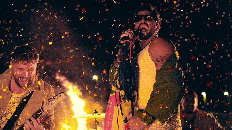 Banda musical en concierto nocturno con fuego y chispas doradas, destacando el cantante con camiseta amarilla y gafas de sol.