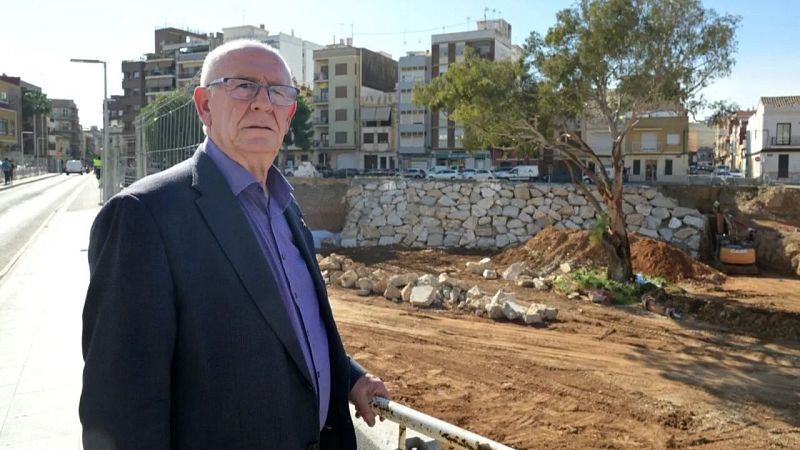 Un hombre con gafas y americana oscura posa en primer plano con un barranco en obras y edificios al fondo.
