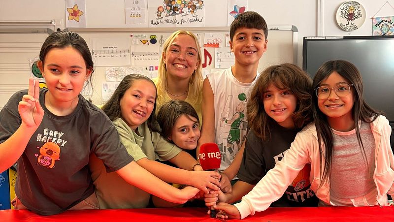 Cinco escolares y una mujer joven sonríen alegremente mientras colocan sus manos sobre un micrófono con el logotipo de RNE en un aula decorada con motivos musicales.