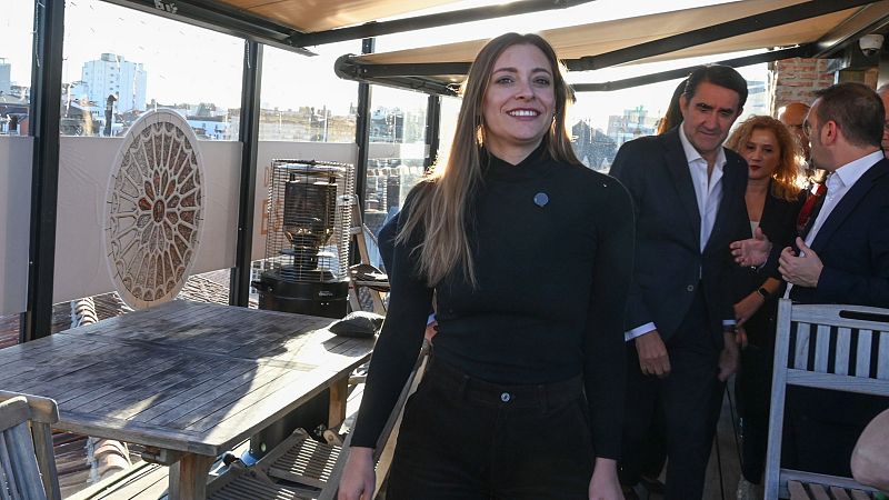 La política Esther Muñoz sonríe mientras camina por una terraza acristalada con estufa exterior y ventanales con vistas a edificios.