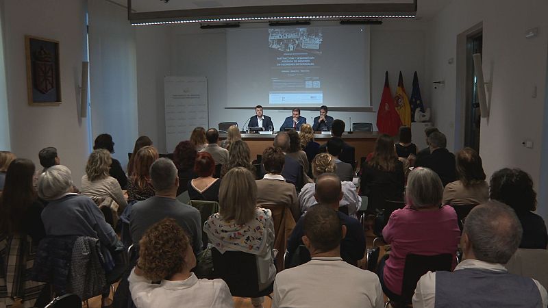Sala de conferencias institucional con público de espaldas, tres ponentes en la mesa presidencial y las banderas de Navarra, España y la Unión Europea.