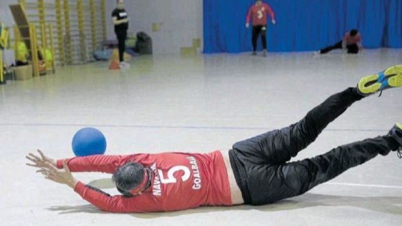 Un deportista con camiseta roja y antifaz protector se estira en el suelo para bloquear un balón azul en un partido de goalball en pista cubierta.