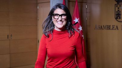 La política Manuela Bergerot sonríe a cámara vistiendo prenda roja y gafas negras ante un fondo institucional con paneles de madera.