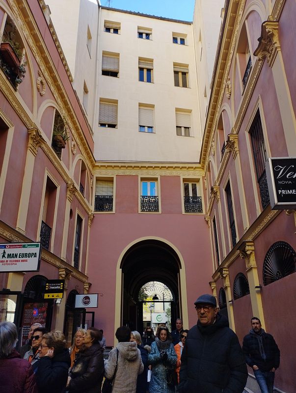 Pasaje peatonal estrecho con edificios clásicos rosa pálido y detalles amarillos.  Se observan comercios y personas caminando.