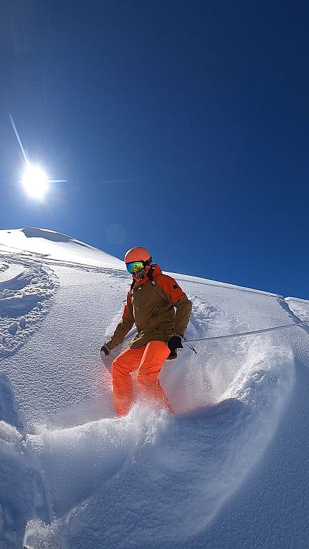 Esquiador con casco naranja y chaqueta marrón baja por una pista nevada, bajo un cielo azul intenso y soleado.