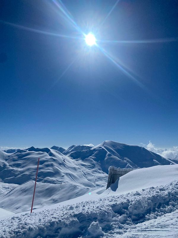 Impresionante paisaje nevado con cielo azul, sol brillante y nieve acumulada. Se aprecian una vara roja y una valla parcialmente cubierta por la nieve.
