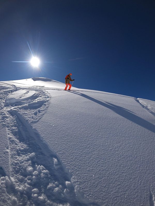 Esquiador en traje naranja sube una ladera nevada, dejando huellas en la nieve polvo bajo un cielo soleado.  Sombra larga proyectada por el sol.