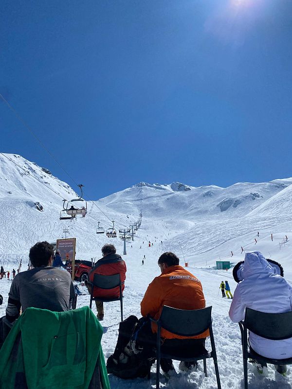 Día soleado en una estación de esquí con personas sentadas observando una pista y un telesilla en funcionamiento. Montañas nevadas y cielo azul.