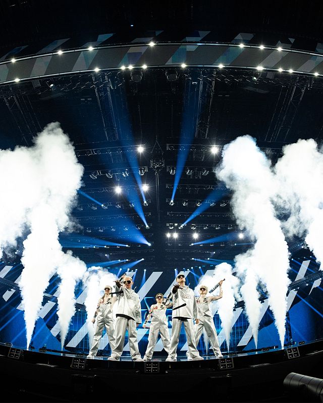 Grupo de 5 personas en escenario con iluminación azul y blanca, humo, y coreografía. Visten monos blancos, gafas y gorras.
