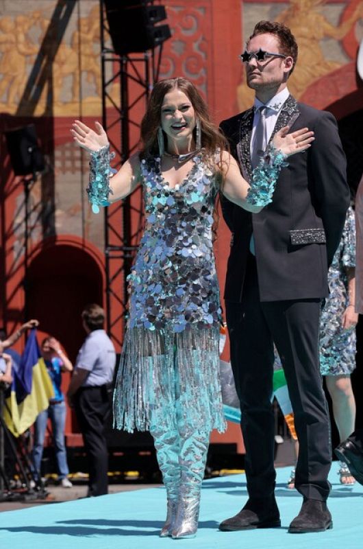 Hombre en traje y mujer con vestido plateado saludan desde una alfombra turquesa con banderas al fondo.