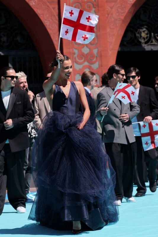 Mujer con vestido azul marino ondea bandera de Georgia en alfombra turquesa.  A su alrededor, grupo de personas con banderas similares.  Posible evento Eurovisión.