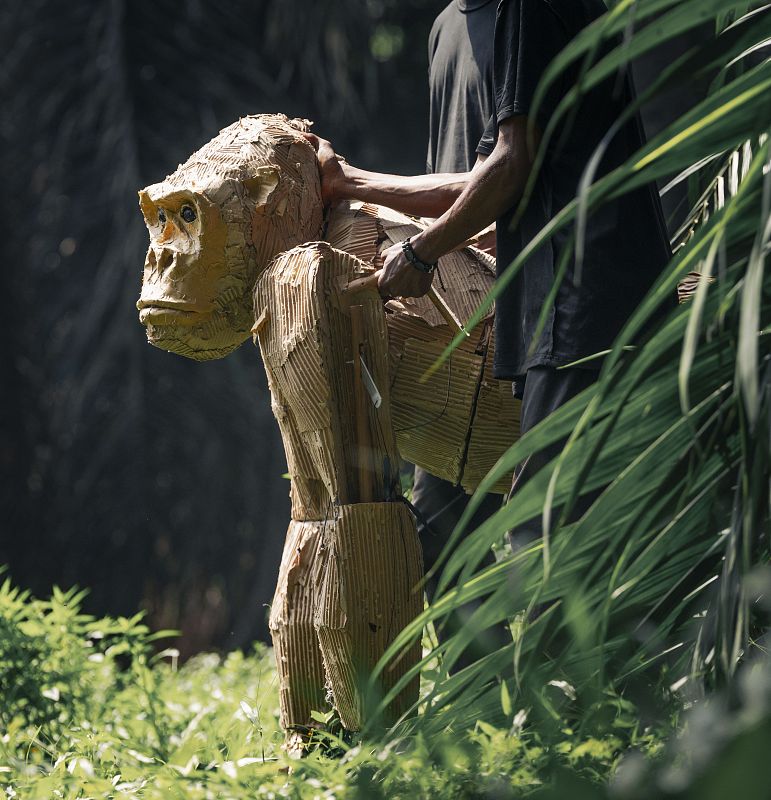 Marioneta de orangután de cartón, sostenida por una persona con camiseta negra, en un jardín botánico con abundante vegetación.