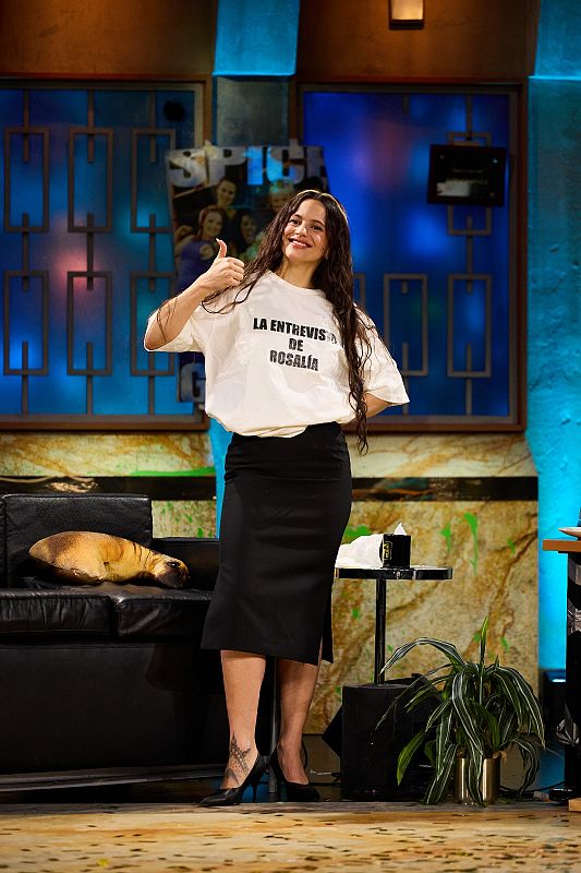 En un set televisivo, una mujer sonríe y hace un gesto de aprobación, luciendo una camiseta con el texto "LA ENTREVISTA DE ROSALÍA", falda negra, tacones y un tatuaje en el tobillo.