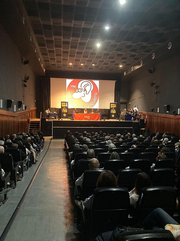 Un auditorio repleto acoge un evento con una pantalla que exhibe el logo de RNE y el lema "No es un día cualquiera", con personas sentadas en el escenario y el público ocupando las butacas.