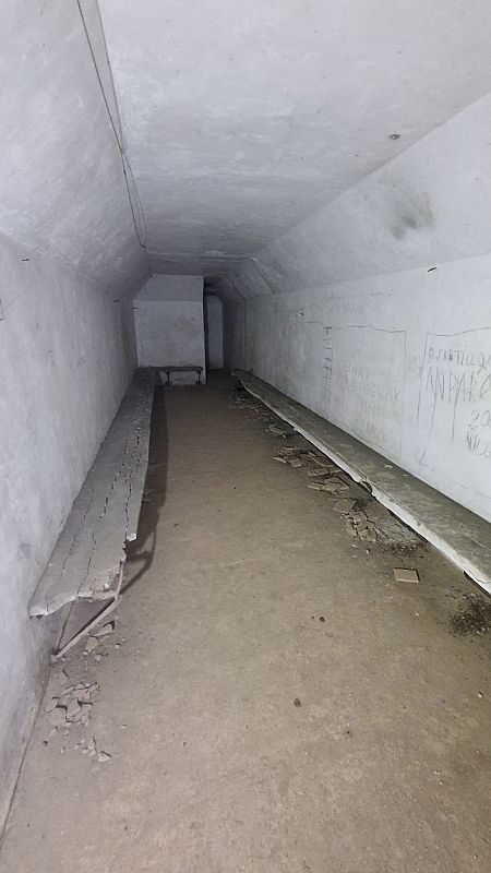 Una sala alargada y estrecha, posiblemente subterránea, con paredes de hormigón y bancos deteriorados. Se observan inscripciones en una pared y el suelo es de tierra o cemento, con restos de material desprendido.