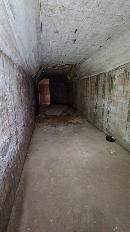 Un túnel subterráneo con paredes de ladrillo y techo de hormigón, iluminado tenuemente, con manchas en el suelo de cemento y una abertura al final.