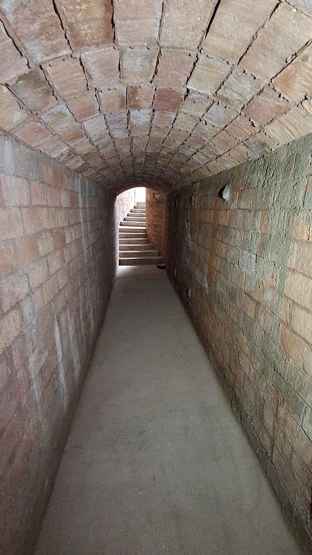 Un túnel de ladrillo con techo abovedado y suelo de cemento, con una escalera al final que conduce a una fuente de luz, creando un ambiente con iluminación tenue.