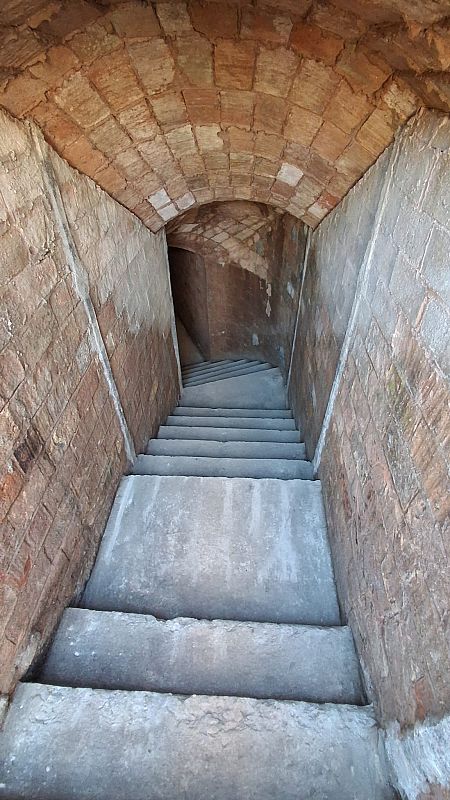 Se observa la entrada a un refugio subterráneo con un pasillo abovedado de ladrillos y escaleras que descienden, creando una sensación de profundidad y protección.