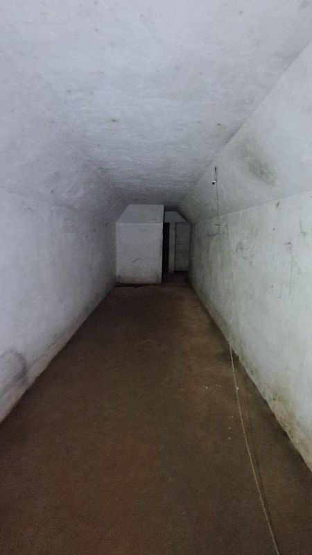 Interior de una sala con paredes y techo abovedado blancos, suelo marrón y una estructura cúbica blanca al fondo con una abertura. Se aprecia desgaste en las paredes y una cuerda colgando.