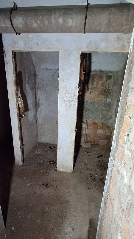 Interior de letrinas abandonadas, con paredes de ladrillo y cemento deterioradas. Se distinguen dos compartimentos y una tubería que atraviesa el techo, con el suelo cubierto de escombros.