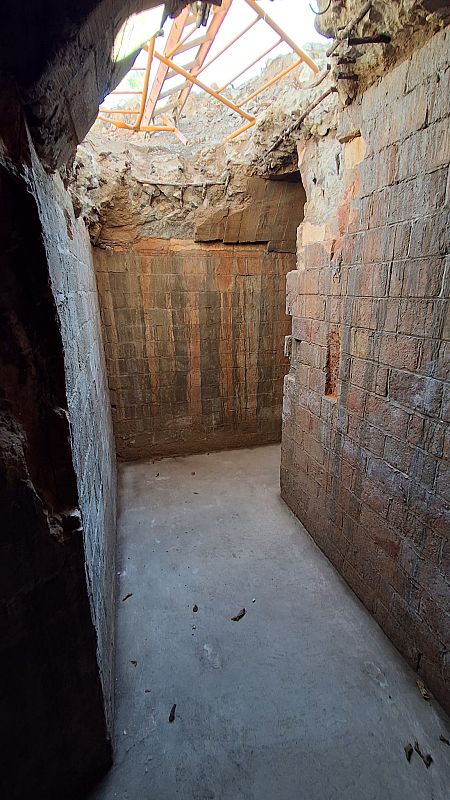 Una esquina subterránea con paredes de ladrillo rojizo deterioradas y suelo de cemento gris. El techo parcialmente destruido expone una estructura metálica y permite el paso de la luz, indicando un espacio abandonado.