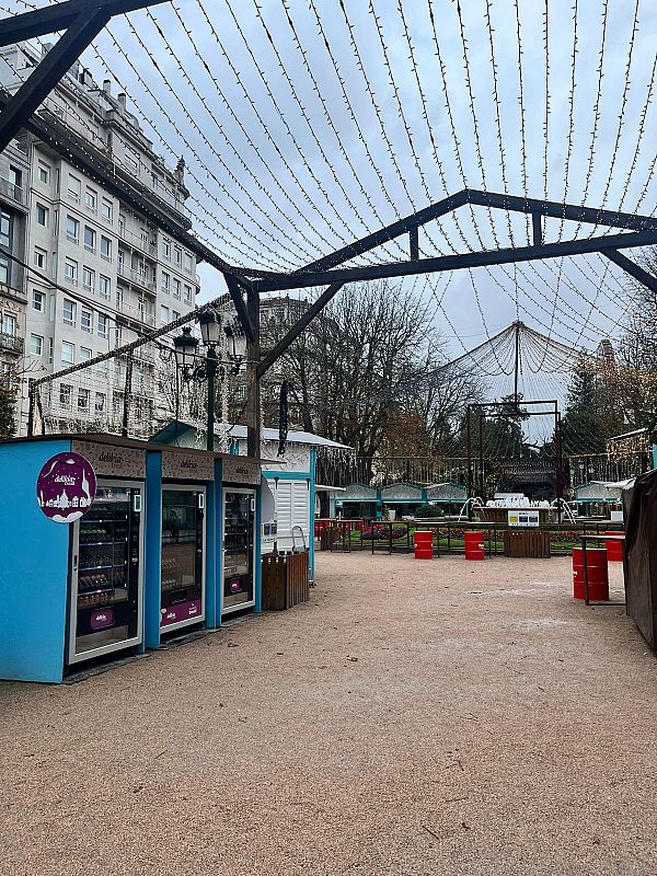 El Cíes Market en Vigo, España, se presenta con una estructura metálica y luces sobre puestos azules de mercado con la marca "delikia", exhibiendo productos. Al fondo, un edificio clásico completa la escena.