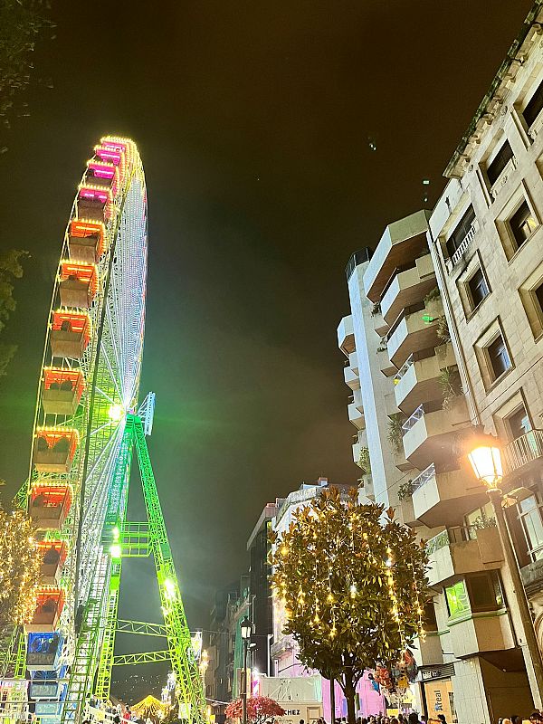 Una noria iluminada con cabinas de colores rojo, azul y naranja se alza en la noche. Luces de colores adornan la estructura de acero blanco, mientras que edificios y una farola se distinguen en el fondo.