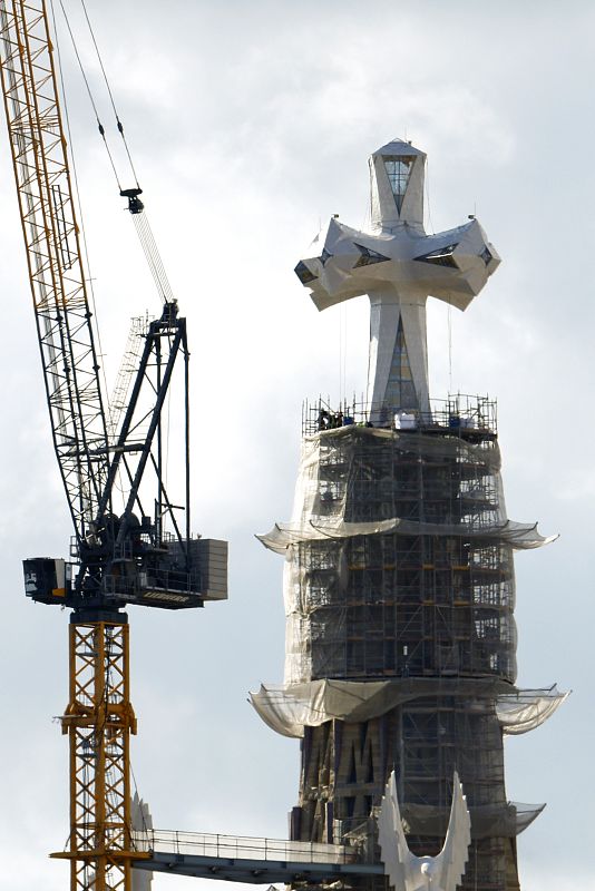 Una vista cercana de la Torre de Jesús en construcción, con una grúa y andamios. En la cima, una gran cruz blanca con diseño facetado y ventanas triangulares.