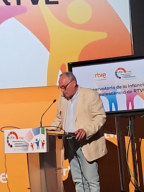 Esteban, con gafas y traje claro, habla en un evento ante un atril con logo de RTVE y "Observatorio de la Infancia y la Adolescencia". Una pantalla al fondo muestra el mismo observatorio.