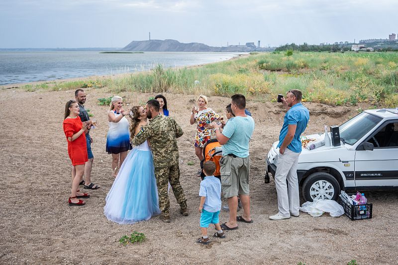 Boda sencilla de dos soldados en una playa, con invitados y un coche cercano.  Se aprecia el mar al fondo.