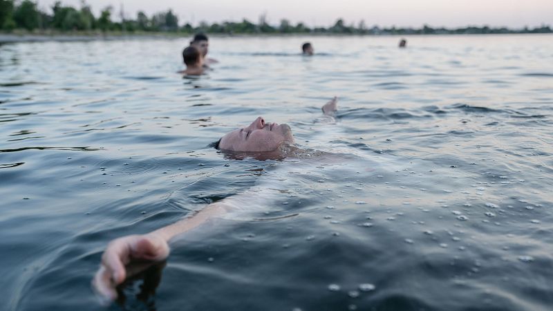 Soldado flotando en un lago, brazos extendidos, ojos cerrados.  Imagen evoca serenidad en medio de la guerra. Fondo borroso con otras figuras en el agua.