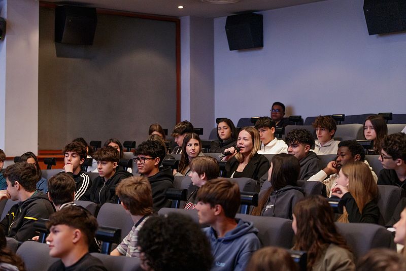 Estudiantes de secundaria escuchan a una compañera que habla por micrófono en una sala de actos durante una gala de bienvenida.  Asientos grises y altavoces en la pared.