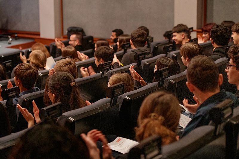 Jóvenes aplauden en un auditorio, algunos con papeles en mano, en una gala con ambiente positivo.