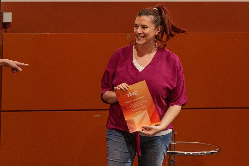 Mujer sonriente con blusa morada y vaqueros, sosteniendo un documento con logo RTVE, es señalada por una mano en un evento con fondo naranja rojizo.