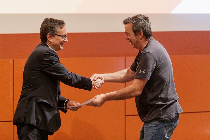 Entrega de un documento doblado entre dos hombres sonrientes en un evento formal con fondo naranja. Uno viste traje y el otro camiseta Under Armour.