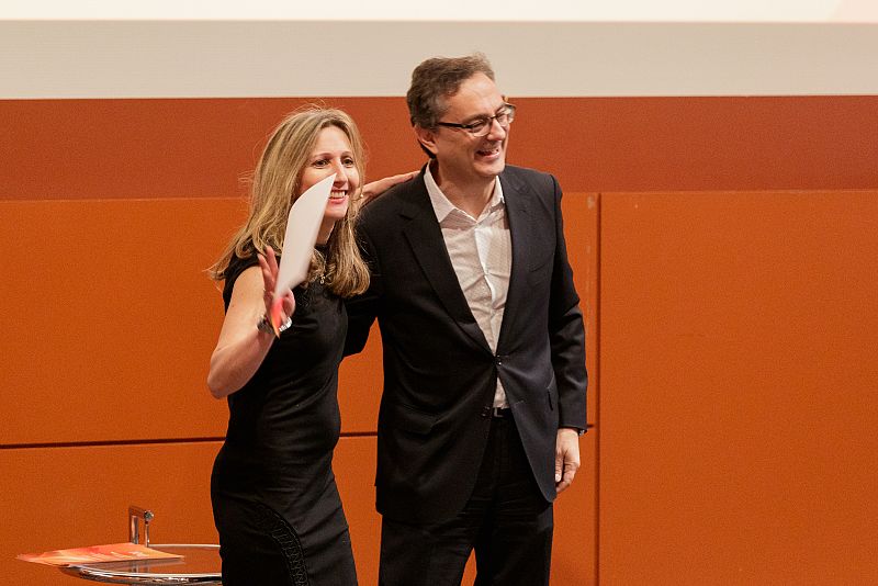 Hombre y mujer sonrientes, él en traje, ella con vestido y papeles. Ambiente positivo, posible evento corporativo o de networking.