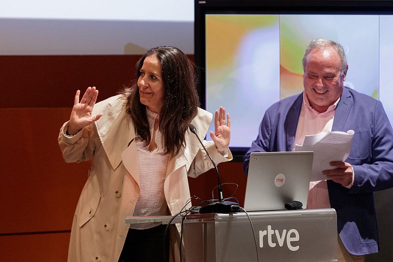 Dos personas gesticulan en un escenario con un atril y el logo de RTVE visible.  Se observa una pantalla grande al fondo.