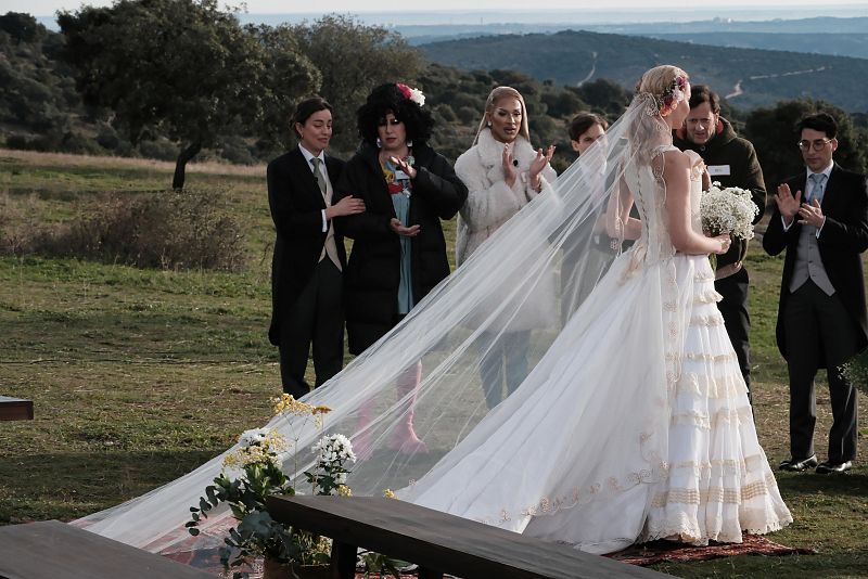 Modelo con vestido de novia blanco, velo y ramo, observada por un grupo de personas en un espacio exterior con vegetación.