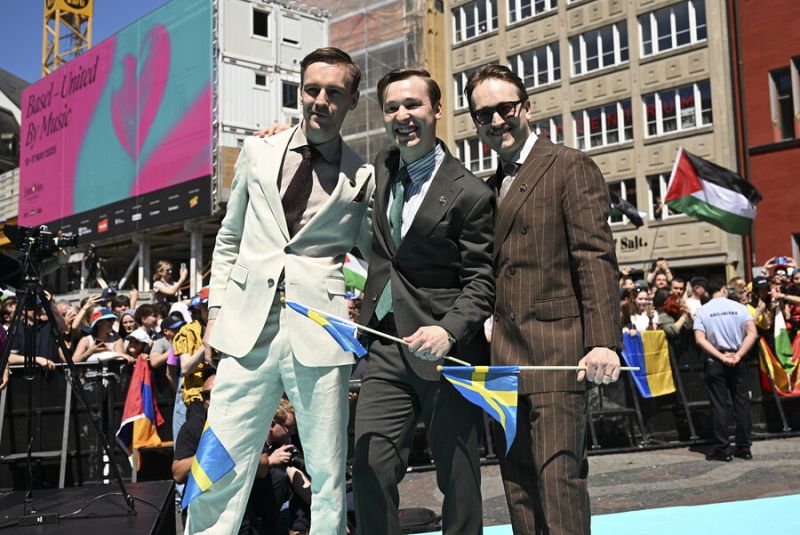 Tres hombres en trajes, con banderas suecas, posan en una alfombra turquesa.  Un cartel de fondo indica 'Basel - United By Music'. Multitud con banderas al fondo.