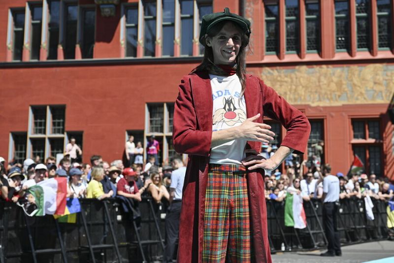 Persona con boina verde, bata granate y camiseta con dibujo, posa en alfombra turquesa con un edificio y multitud de fondo.